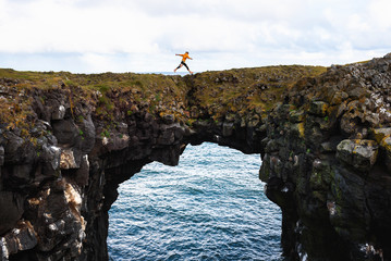 Tourist jumps over a natural rock bridge in Arnarstapi, Iceland