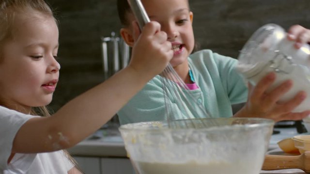 Medium shot of cute little girl adding flour into bowl with dough while her sister stirring it with whisk while learning to cook together