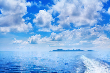 White cumulus clouds in blue sky over sea landscape, many clouds above ocean water panorama, ship trail, island on horizon line, beautiful tropical sunny summer seascape panoramic view, cloudy weather