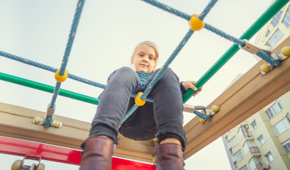 Fototapeta premium A charming little girl in casual clothes sits on the grid and looks at camera on playground in the urban district.