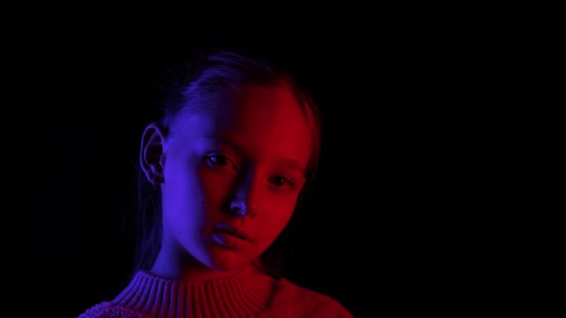 Young girl looking up on black background in dark studio with blue and red lighting. Portrait girl teenager posing front camera in black studio with neon lighting background