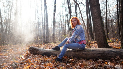 Fototapeta premium Woman sitting on log in autumn forest. Young woman with red hair in outwear sitting on fallen log in rural forest looking away in sunlight