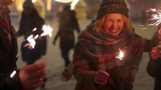 Cheerful friends have fun waving with sparklers in hands. People partying on winter night with snowfall, lamp garlands on backdrop at Christmas market square in slow motion. New Year, Birthday party.
