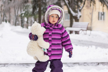 A little girl in a hat, hat and boots walks along the street with a teddy bear in the winter season