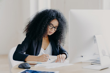 Photo of serious Afro woman writes in papers, sits at table with modern computer, creats article in...