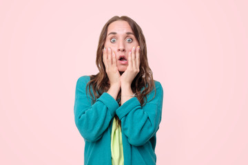 Surprised woman with opened mouth and big eyes holding hands near face and looking happy on pink background. Studio shot