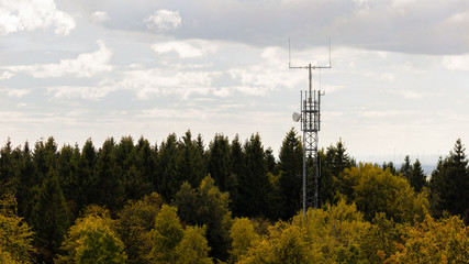 Obraz premium A mobile radio mast in the middle of a autumn forest on a mountain in Germany. The leaves are partly yellow. In the sky are interesting and beautiful clouds.
