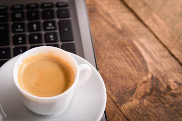 Cup of coffee on a laptop keyboard. Work place modern. Laptop (notebook) with cup of coffee and notepad with pen on old wooden table.