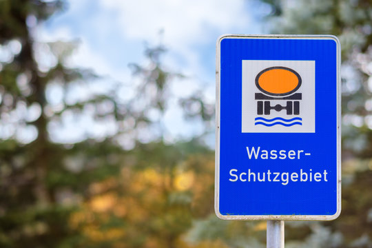 In a rural area with forest is a rectangular blue warning sign. It is a Water Reserve. A tank truck is pictured on the sign. Translation: water protection area