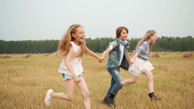 Happy teenager friends holding hands running on harvesting field in village. Carefree teenager boy and girls running on haystack background on countryside field