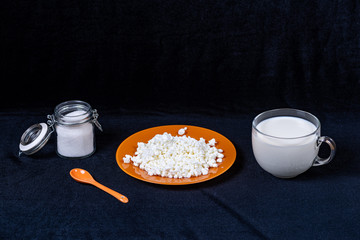 Glass jar with sugar, orange porcelain spoon, plate with cottage cheese and glass mug with milk on a dark background