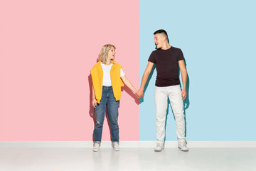 Obraz premium Young emotional man and woman in bright casual clothes posing on pink and blue background. Concept of human emotions, facial expession, relations, ad. Beautiful caucasian couple holding hands and