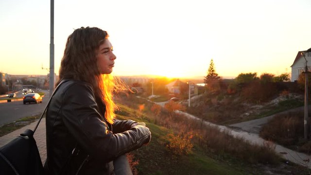 A young beautiful woman in a leather jacket with a black backpack stands thoughtfully on the bridge against the background of the urban district.
