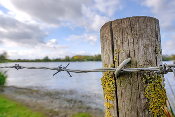 Close-up, shallow focus of barbed wire seen attached to a weathered, wooden post, seen at a nature reserve lake. The lake contains many rare migratory birds from the UK and beyond.