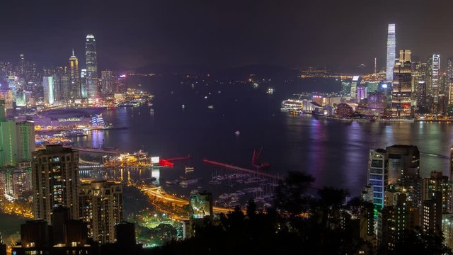 Timelapse Hong Kong city highrise buildings surround harbour with sailing boats and city reflection on dark water against hill silhouettes at night zoom out