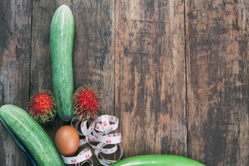 A concept cucumber made into a penis with a tape measure beside it on a dark brown wood floor.