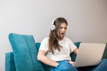 young beautiful woman in headphones listening to her favourite music from laptop and playing video game on the sofa in the living room looking angry
