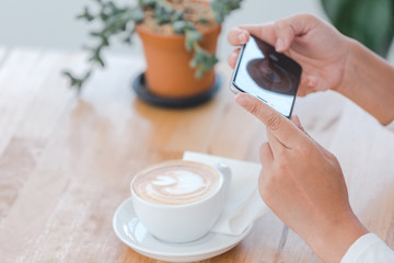 A good girl uses a camera from a smartphone to capture a white coffee cup resting on a brown wooden table.