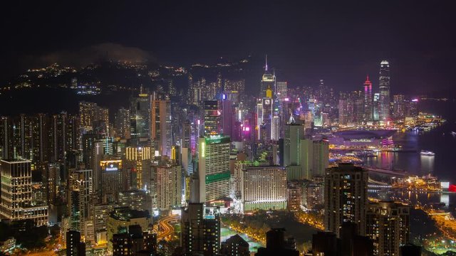 Timelapse flashing advertisements on Hong Kong Central and Western districts skyscrapers near large harbour against hill silhouettes at night zoom out