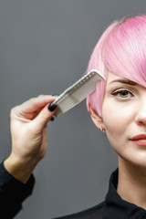 Hand of professional hairdresser is combing with a comb short pink hair of beautiful young woman on gray background. Close up. Copy space.