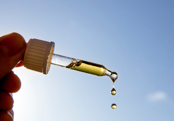 Hand holding dropper pipette with nice golden liquid D-vitamin against sun and blue sky on sunny...