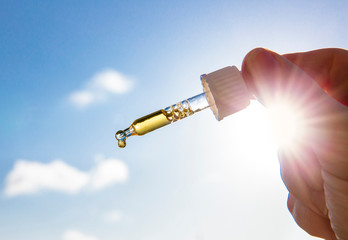 Hand holding dropper pipette with nice golden liquid D-vitamin against sun and blue sky on sunny...