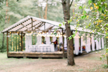 Blurred photo of wooden tent restaurant with wooden tables and chairs., decorated with light bulbs Vintage design of the restaurant in the forest in summer. Wedding reception in the open air.