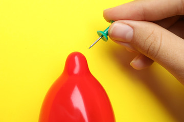 Woman piercing inflated condom with pin on yellow background, closeup. Safe sex