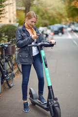 Young woman with electric kick scooter in the street of the city, looking at her phone and smiling. Public transportation renting service, modern urban ecological zero emission transport concept. © Lars Zahner