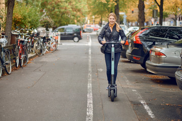 Young happy blond woman riding an electric scooter in the city, smiling at the camera, in autumn