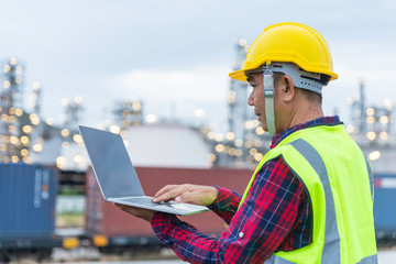 Male foreman in yellow safety helmet is looking to The computer in front of blurred background of refinery station