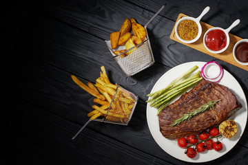 grilled steak on a plate with asparagus and tomatoes, French fries and country potatoes. Three different sauces: ketchup, mustard and barbecue on a dark wooden background, top view, horizontal photo
