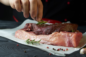 Chef salts steak with herbs on paper. Preparing fresh beef or pork. Horizontal photo with a dark black background.