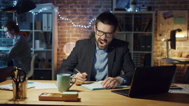 Tired businessman in suit is yawning working in office writing in notebook sitting at desk alone feeling exhausted. Workaholics, workplace and people concept.