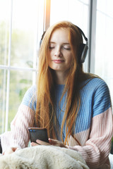 pretty young red haired woman with headphones listening to music at home, relaxing, enjoying songs