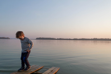 Fototapeta premium Little kid boy on a wooden pier on a lake, a river looks into the distance at the horizon