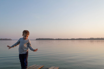 Little kid boy on a wooden pier on a lake, a river looks into the distance at the horizon