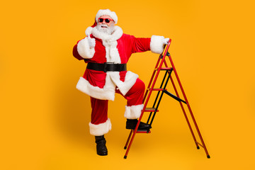 Full size photo of funky white beard hair santa claus in red hat climb ladder decorates christmas tree deliver wish dream gifts show thumb up approve x-mas celebration isolated yellow color background © deagreez