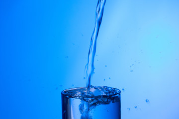 Chorro de agua llenando y rebosando un vaso de agua de cristal o plástico transparente sobre fondo azul