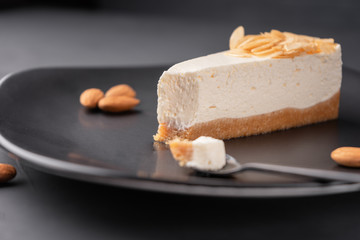Cheesecake on a black plate among the almonds, slice incision in the spoon, close-up.