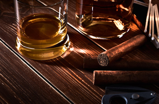 Still life with glass and bottle of alcohol, whiskey or brandy or rum. Three cuban cigars, cutter and matches on old wooden table top