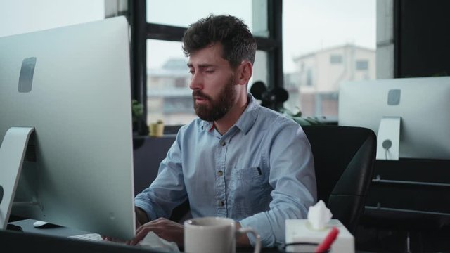 Poor tired office worker feeling sick sneezing allergy with paper tissues blowing his nose while working at desktop computer in the office.