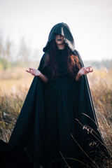 A girl in a black dress, a cloak with a hood. It stands in a high dry grass in the field against...