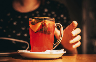 girls hand reaches for a cup with fruit tea, slices of lemon, orange and mint. A cup of tea is on a wooden table in a cafe. The concept of a cozy time in the autumn.