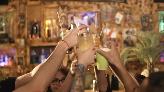 Handheld shot of group of cheerful young friend of different ethnicities dancing at party and clinking glasses with cocktails at bar or nightclub