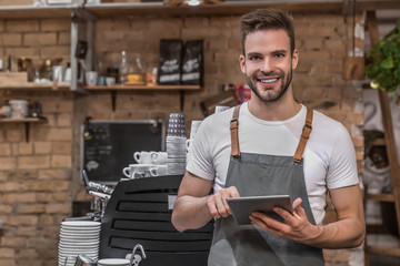 Smiling young entrepreneur wearing an apron near counter of his cafe and using digital tablet