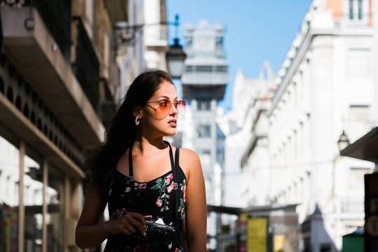 Confident beautiful woman in summer dress holding a photo camera while standing on scenic sunny city street in Lisbon, Portugal