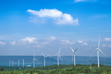 The big white wind turbine for spinning electricity sees large mountains far away, with clouds and blue skies above the mountains.