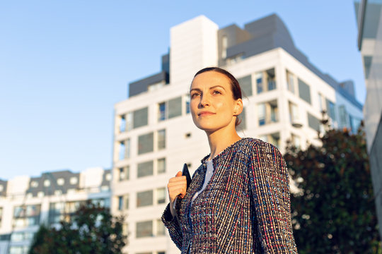 Low angle of serious female manager in elegant outfit leaning on railing and looking away while standing outside modern office building