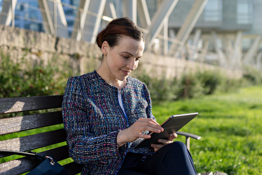 Serious female entrepreneur in elegant jacket frowning and using tablet while sitting on bench on sunny day in city park